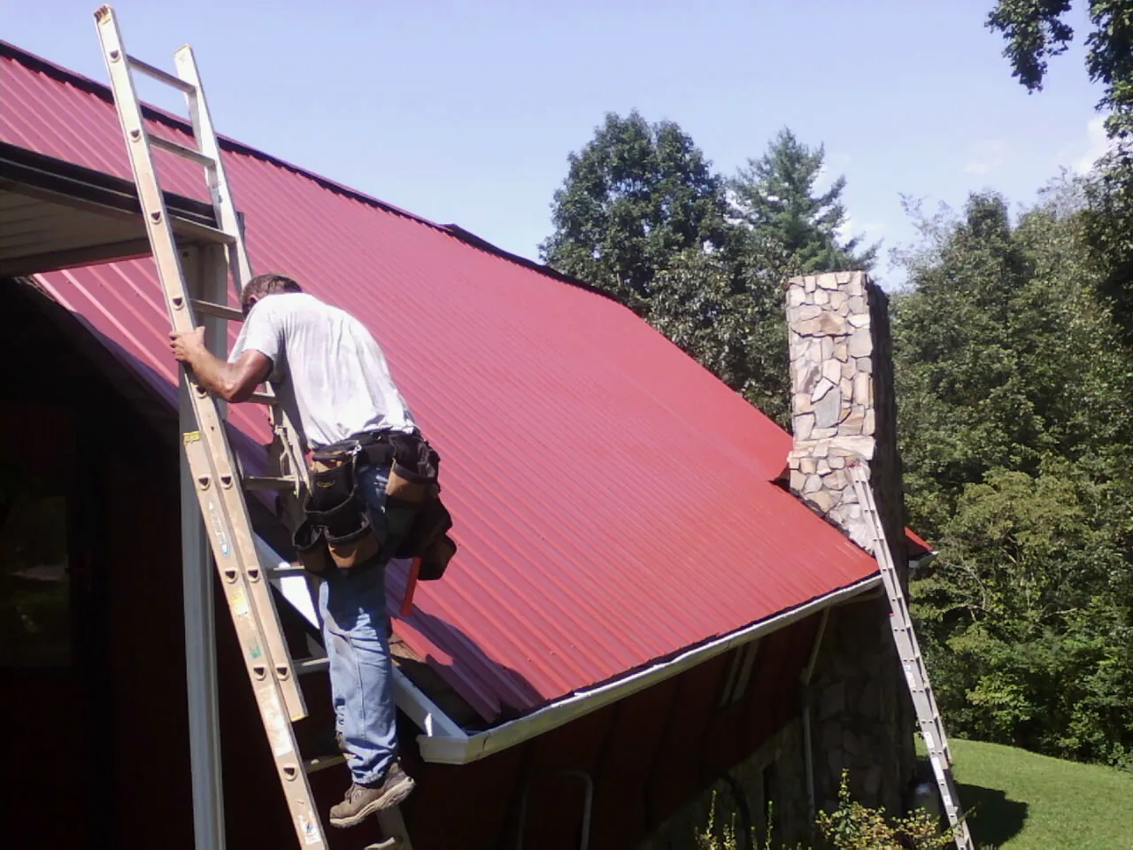 Roofer on a ladder beside a red metal roof and stone chimney — Soffit & Fascia Repair by Roofing Brothers in Hiawassee